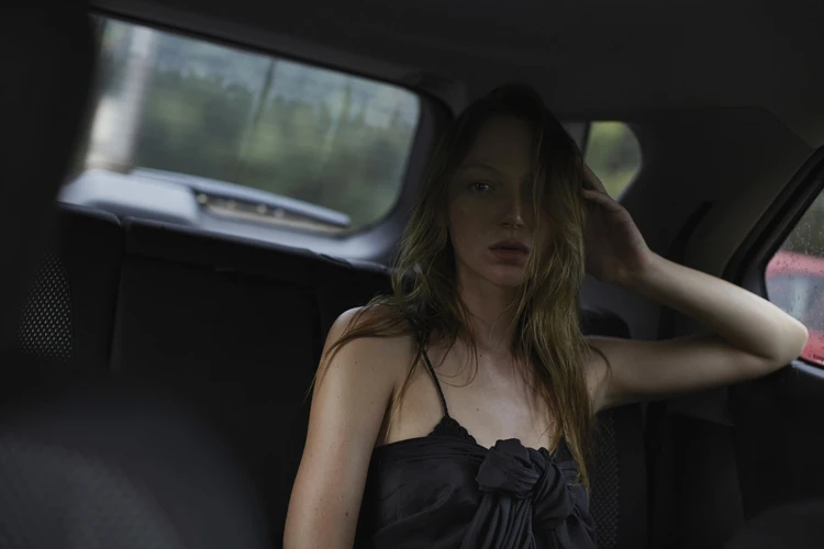 Young woman with long brown hair sits in the backseat of a car, wearing a black spaghetti strap top and looking at the camera.