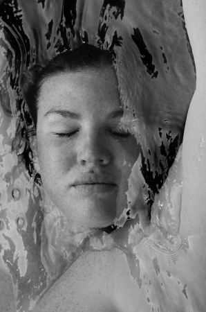 Monochrome portrait of a woman submerged in water with eyes closed and water rippling across her face.
