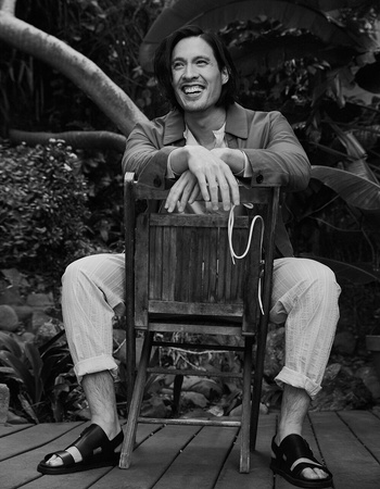 Monochrome portrait of a smiling man with long hair seated backward on a wooden chair outdoors amidst lush foliage.