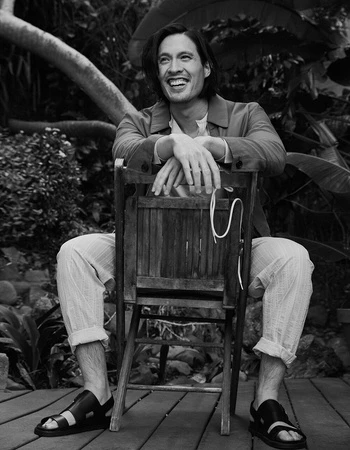 Monochrome portrait of a smiling man with long hair seated backward on a wooden chair outdoors amidst lush foliage.