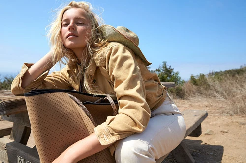 Blonde woman wearing tan jacket and white pants leaning on wooden picnic table holding large perforated tan tote bag
