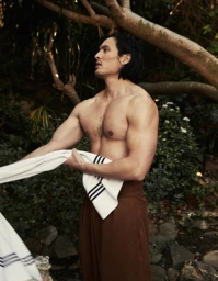 Shirtless man with dark hair holding a white towel with black stripes wearing brown trousers outdoors in a lush tropical garden