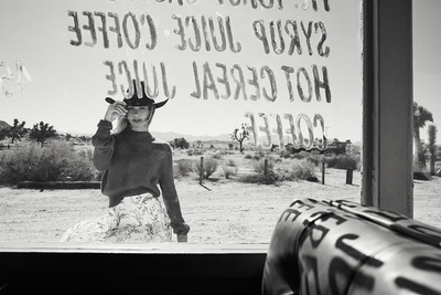Black and white portrait of a woman wearing a cowboy hat and sweater viewed through a glass window with painted text