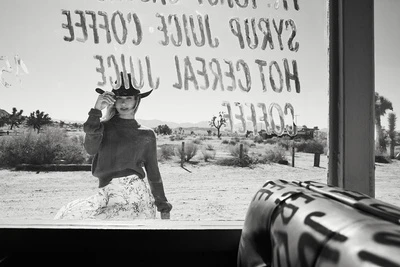 Black and white portrait of a woman wearing a cowboy hat and sweater viewed through a glass window with painted text