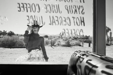 Black and white portrait of a woman wearing a cowboy hat and sweater viewed through a glass window with painted text