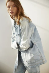 A red-haired female model wearing a light blue denim jacket and jeans poses against a neutral wall indoors.