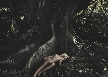 Woman lying down amongst fallen leaves at the base of a massive tree trunk with exposed roots