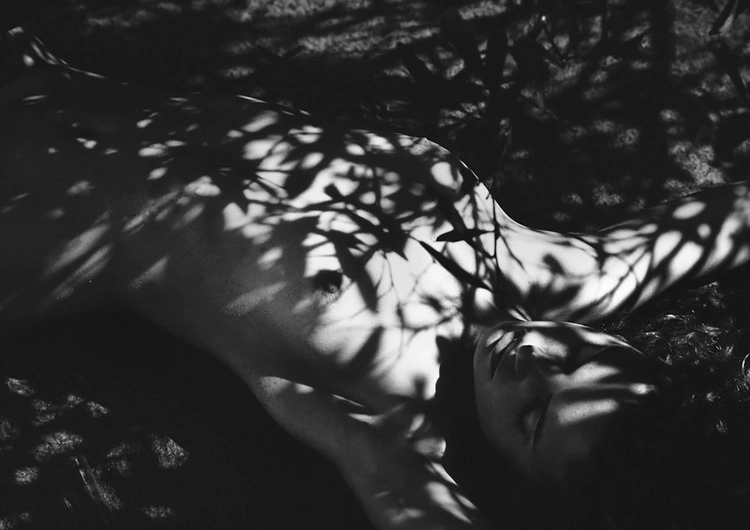 Monochrome image of a person reclining under harsh dappled foliage shadows creating high-contrast patterns across the body.