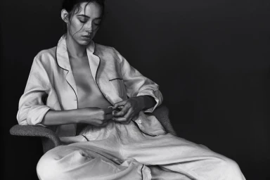 Monochrome portrait of a woman seated in a chair wearing light-colored pajama style button-up shirt and trousers by Sylve Colless.