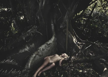 A woman lying on the ground amidst fallen leaves at the base of a massive tree root system in a dark forest.