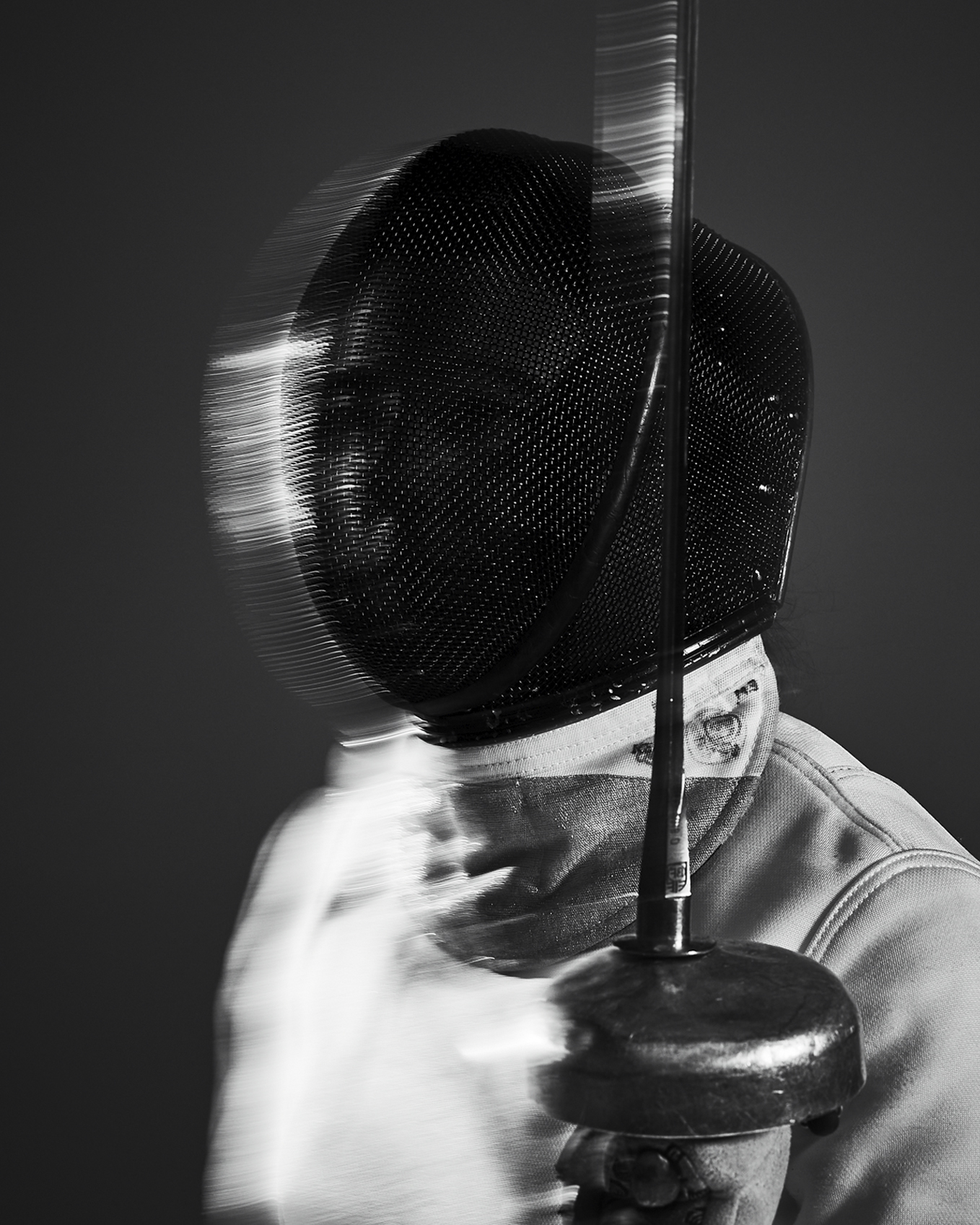 Monochromatic shot of fencer in mask and plastron holding foil. Motion blur suggests swift movement in indoor fencing match.