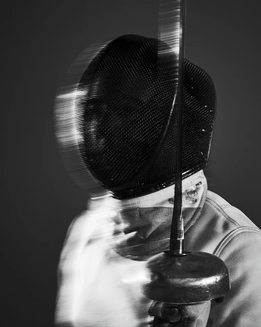 Monochromatic shot of fencer in mask and plastron holding foil. Motion blur suggests swift movement in indoor fencing match.