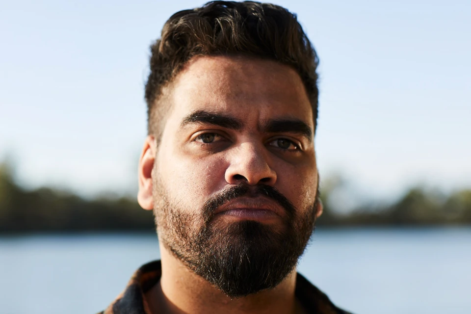 Close-up portrait of a dark-haired adult male with a beard, looking directly at the camera against a blurred outdoor background.