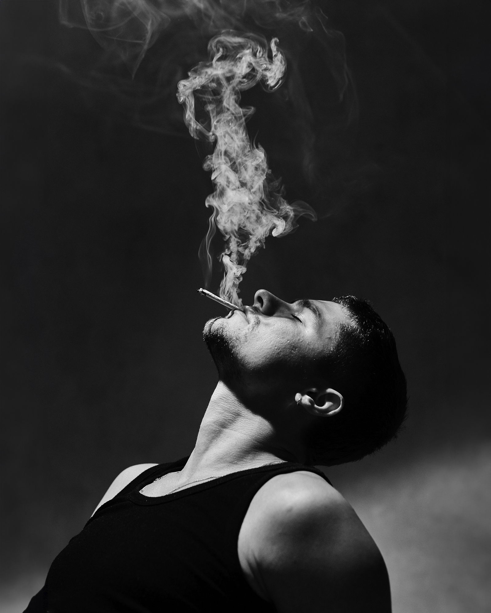 Monochrome portrait of a man smoking, with smoke rising in the air against a dark background. The subject has a serious expression.