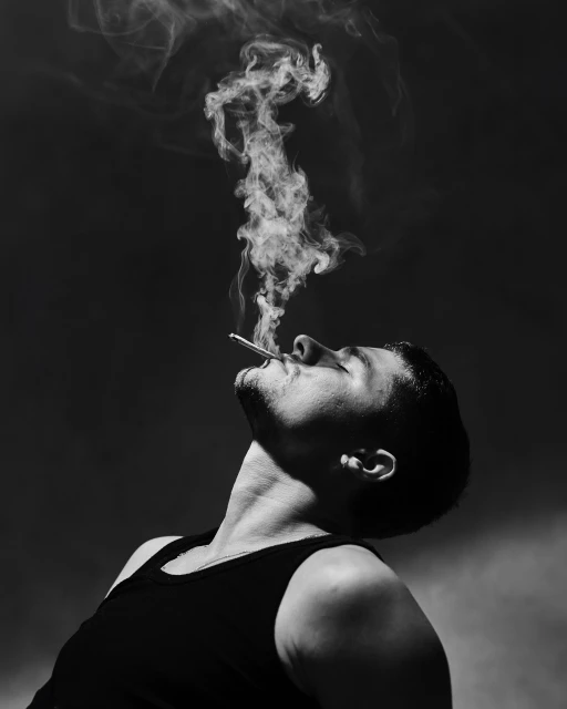 Monochrome portrait of a man smoking, with smoke rising in the air against a dark background. The subject has a serious expression.