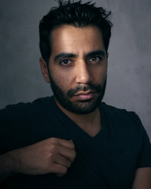 Portrait of a man with dark hair and a beard, looking directly at the camera with a serious expression against a grey textured background.