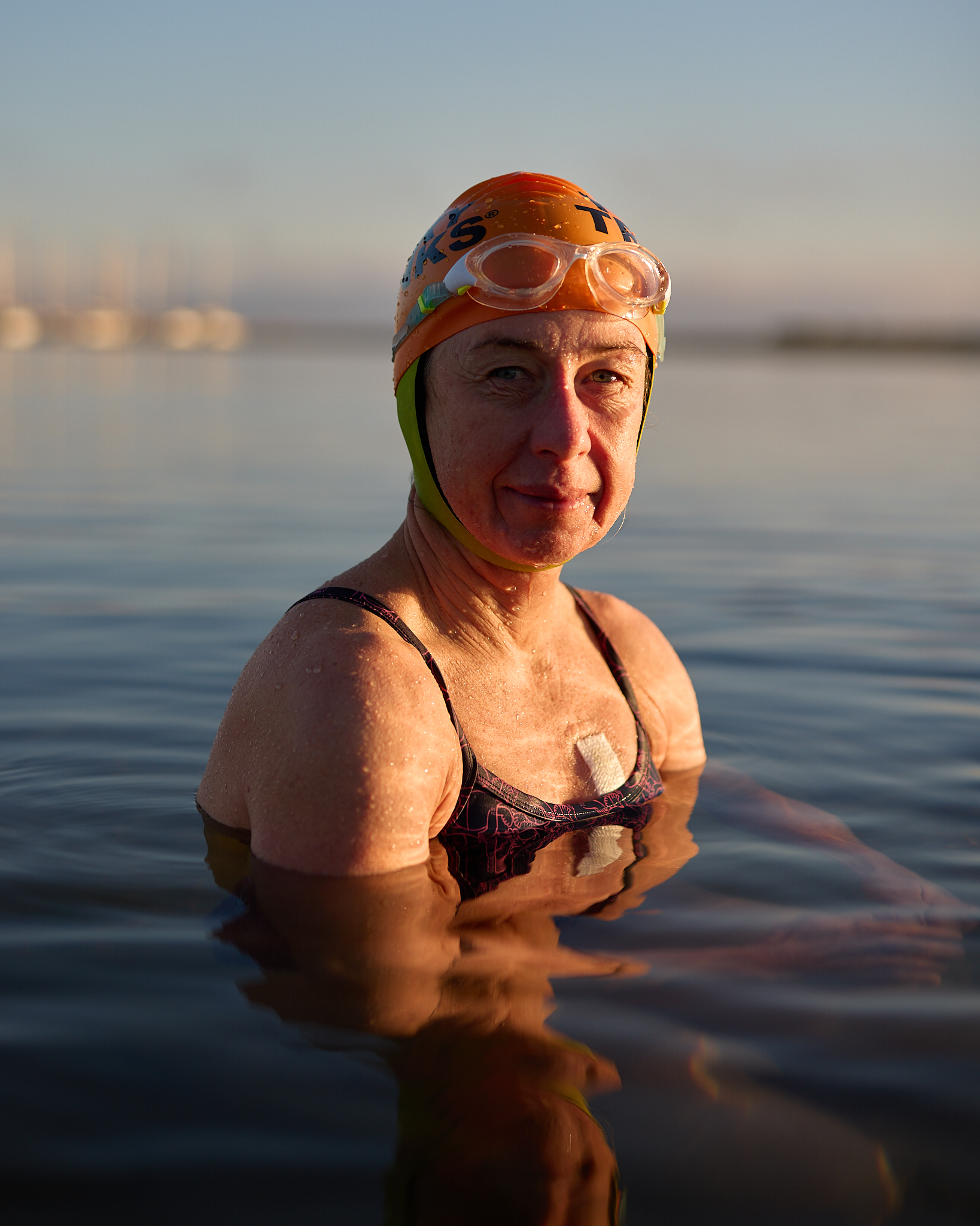 Woman swimmer in outdoor water wearing swim cap, goggles, and bathing suit; water reflects light, creating a serene atmosphere.