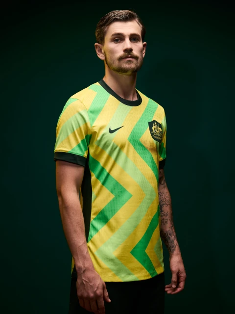 Adult male in a green and yellow chevron patterned soccer jersey with the Australian national team logo on a dark green background.