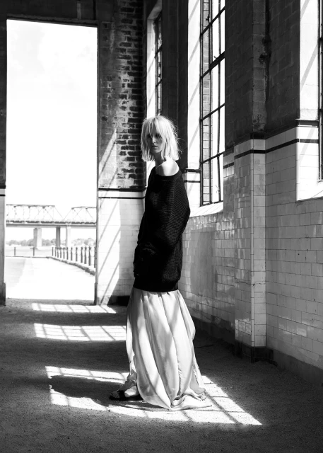 Black and white fashion shot: blonde woman in off-shoulder sweater and skirt poses in derelict building with long shadows.