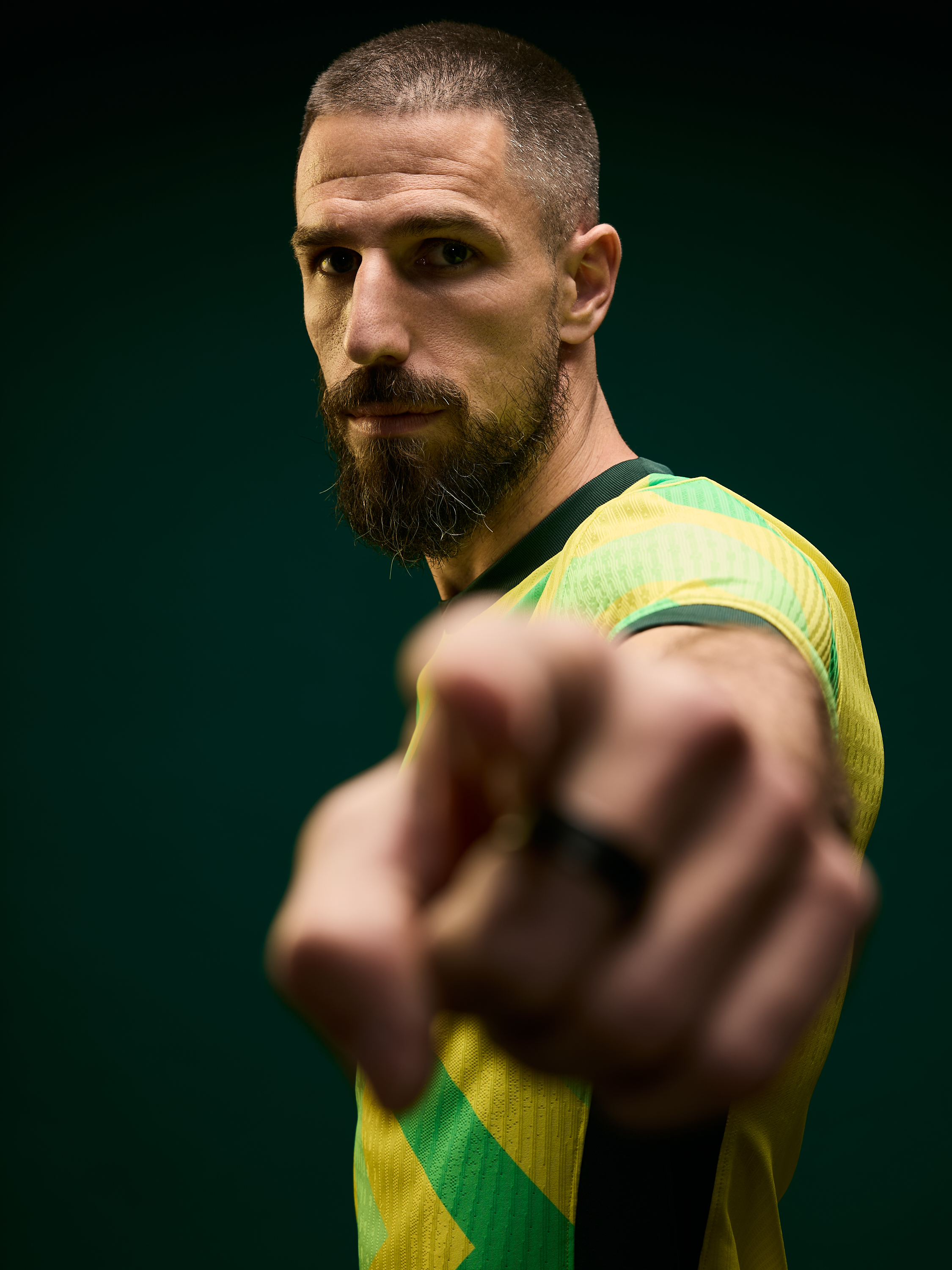 Portrait of a bearded man in a yellow and green sports jersey pointing at the viewer against a dark green background in a studio setting.
