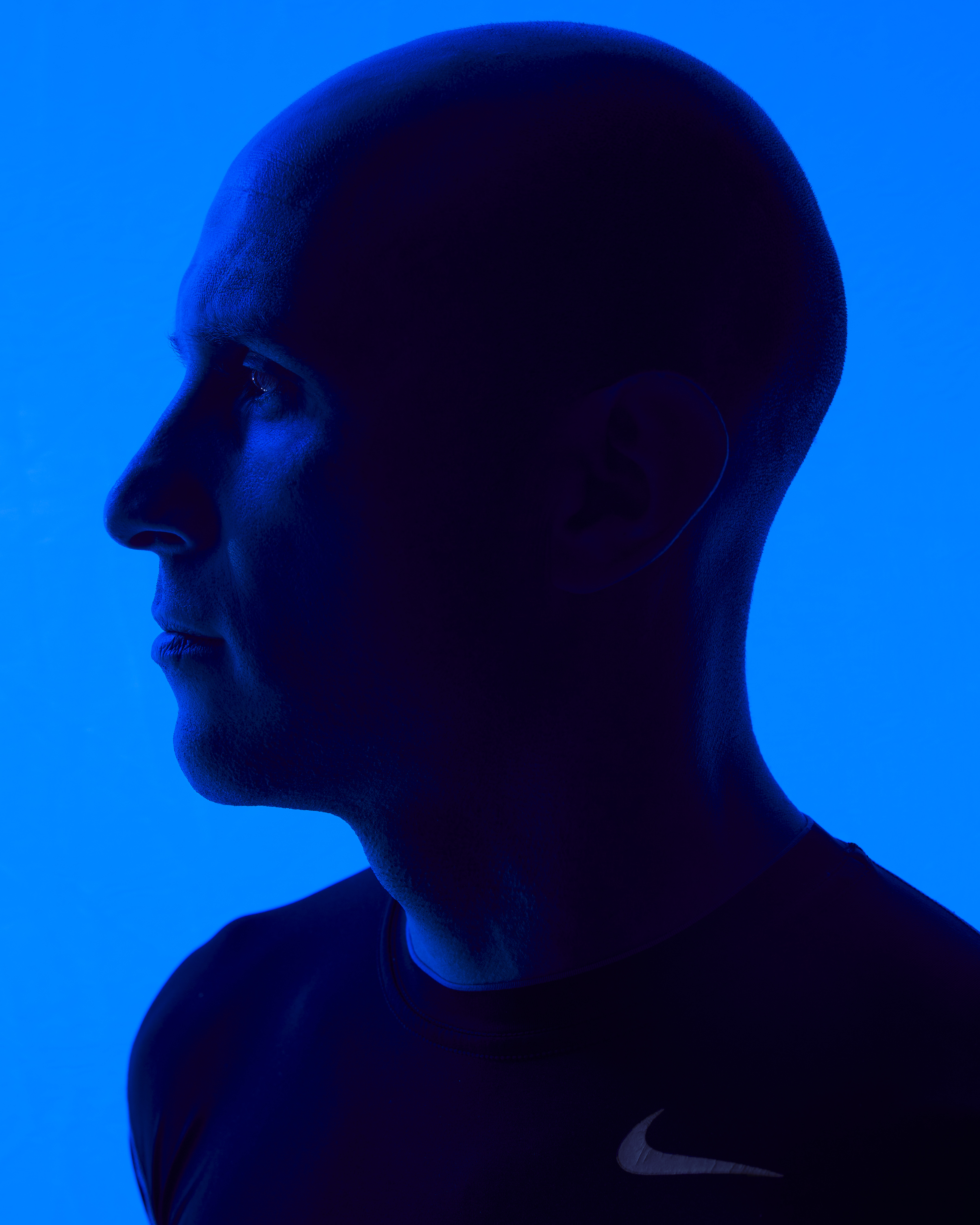 Profile of bald adult male in dark athletic wear in blue rim lighting against a solid blue background in a studio setting.