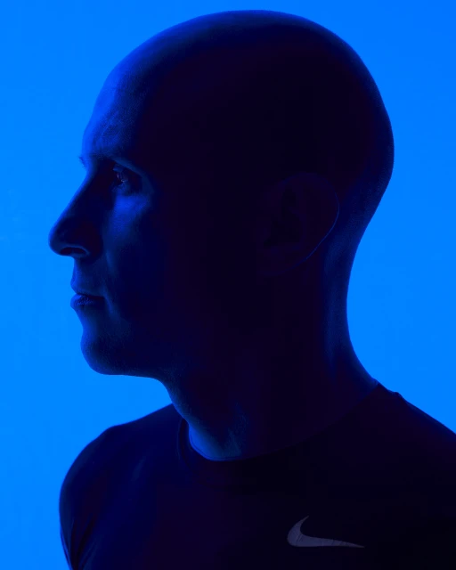 Profile of bald adult male in dark athletic wear in blue rim lighting against a solid blue background in a studio setting.