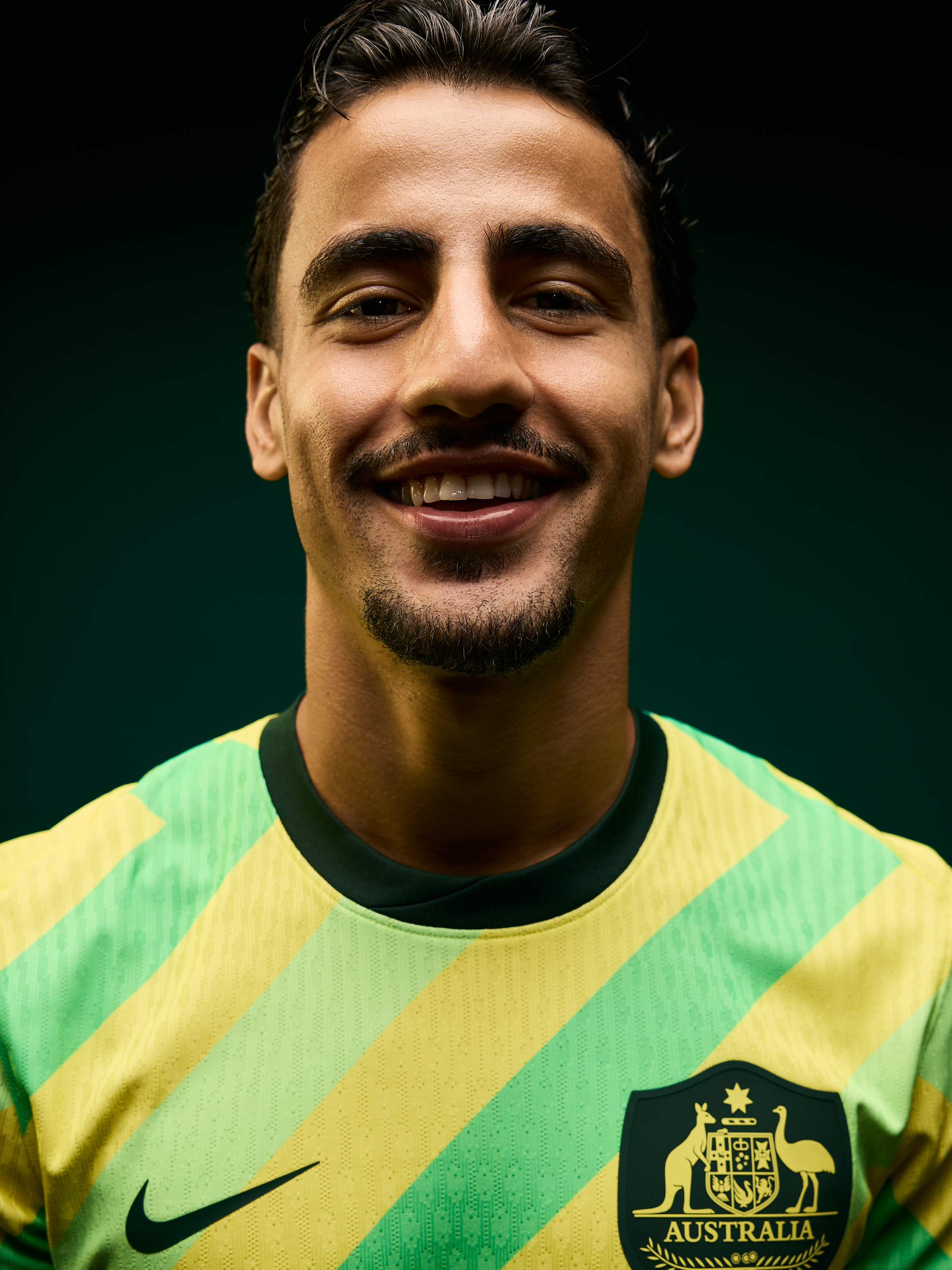 Close-up of smiling man in green and yellow Australia soccer jersey against a dark green backdrop, showcasing team pride.