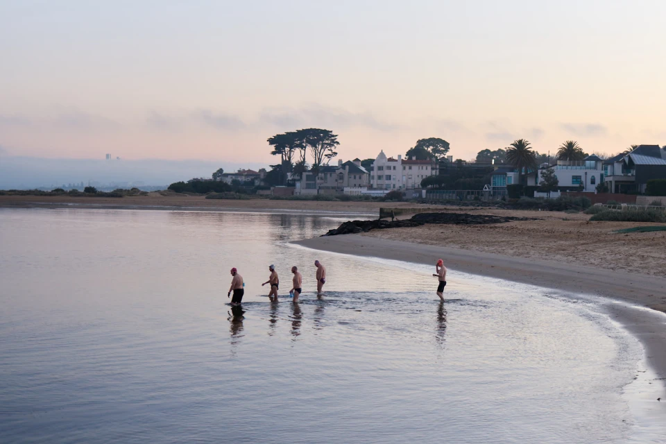 Group of adults in swimwear wading into calm ocean water at sunrise near a sandy beach with coastal houses and trees in the background.