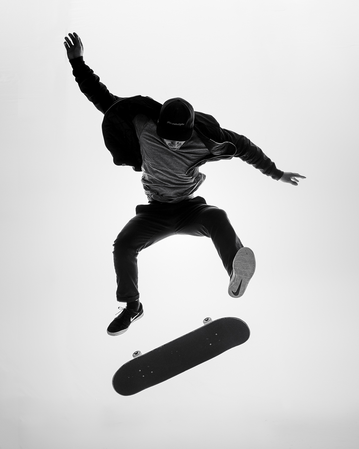 Black and white action shot of a skateboarder in mid-air, skateboard floating below, against a bright white background.