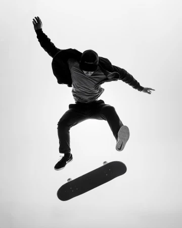 Black and white action shot of a skateboarder in mid-air, skateboard floating below, against a bright white background.