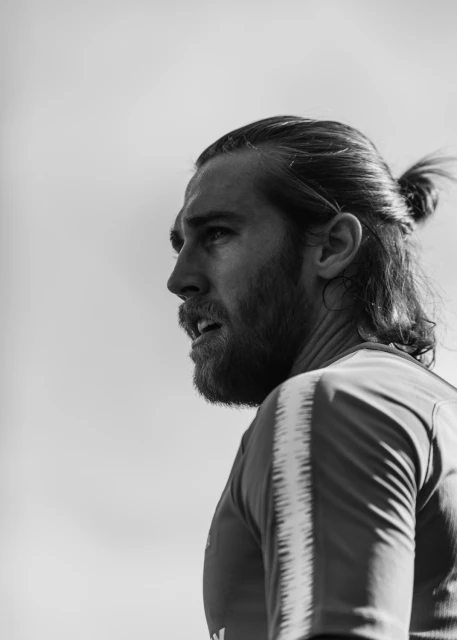 Black and white portrait of a bearded man with long hair in a topknot looking to the left in an outdoor setting with sky background.