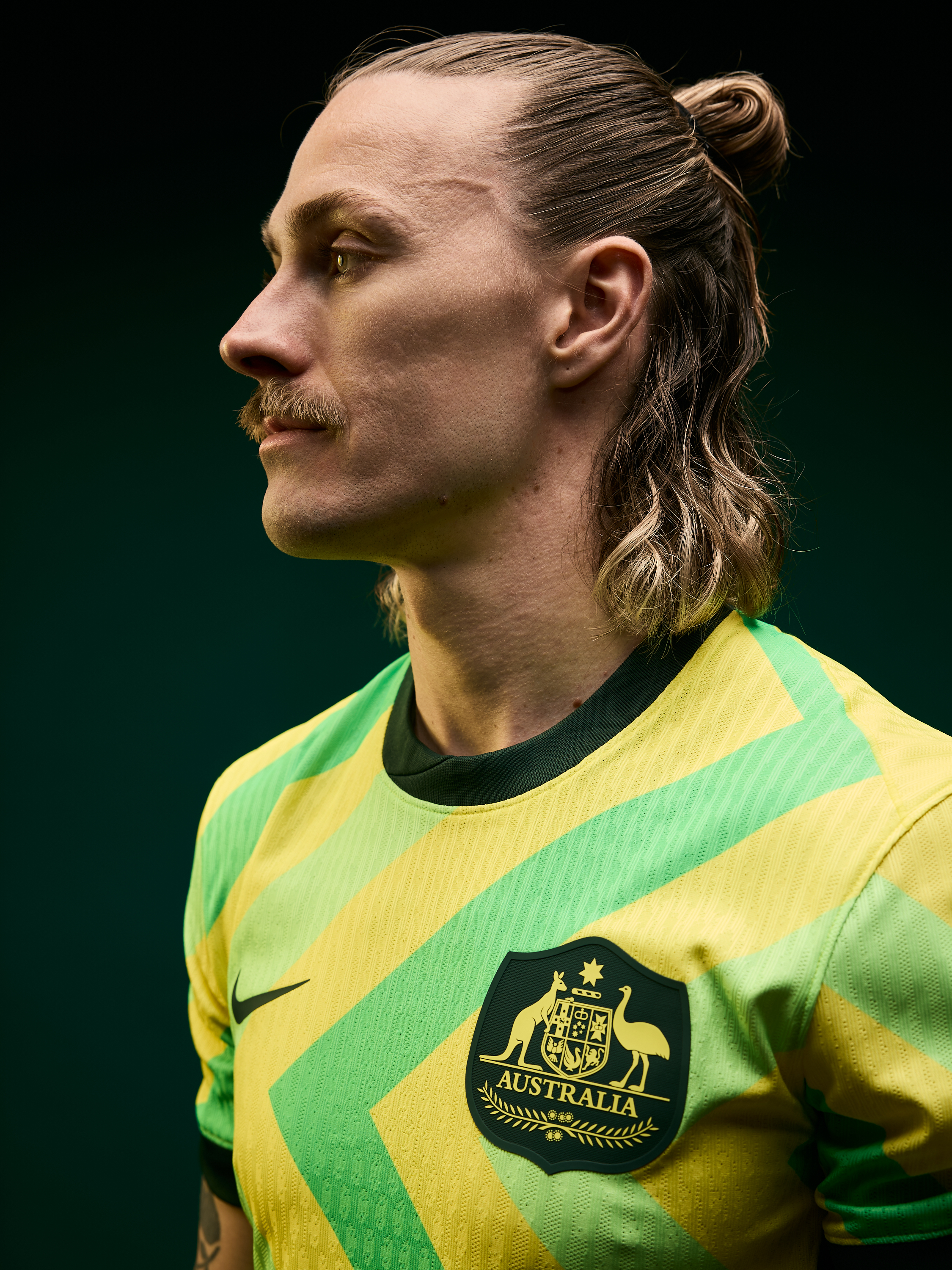 Profile view of an adult male with blonde hair, styled in a man bun, wearing an Australian soccer jersey against a dark green background.