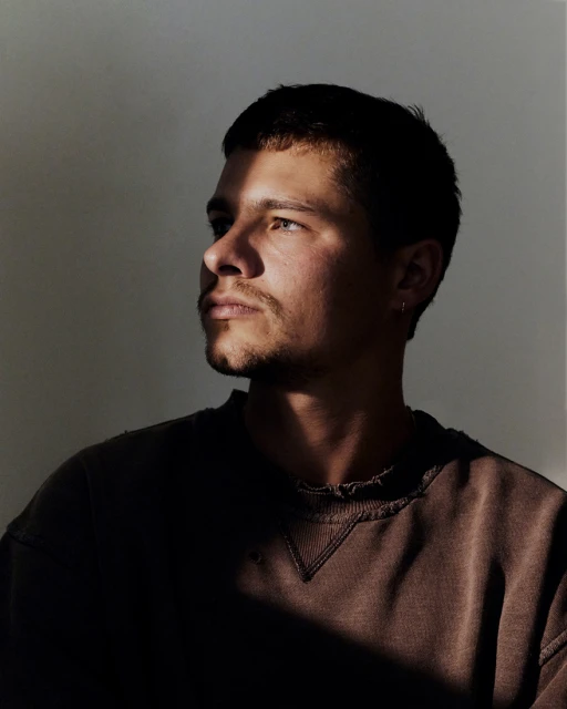 Close-up portrait of a dark-haired man with a beard wearing a brown sweatshirt in soft, directional light against a neutral backdrop.