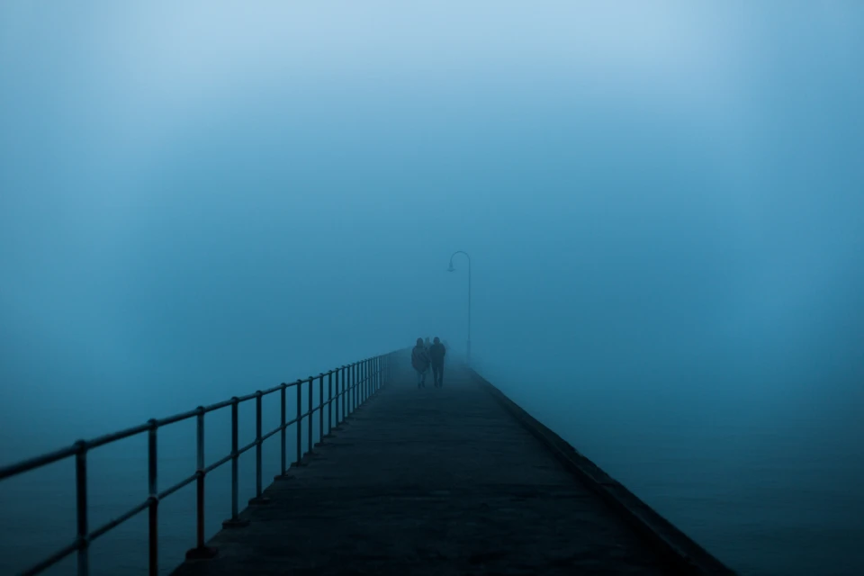 Two people walk into the fog on a pier with metal railings. The heavy fog obscures the water and sky in a monotone blue color.