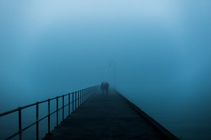 Two people walk into the fog on a pier with metal railings. The heavy fog obscures the water and sky in a monotone blue color.