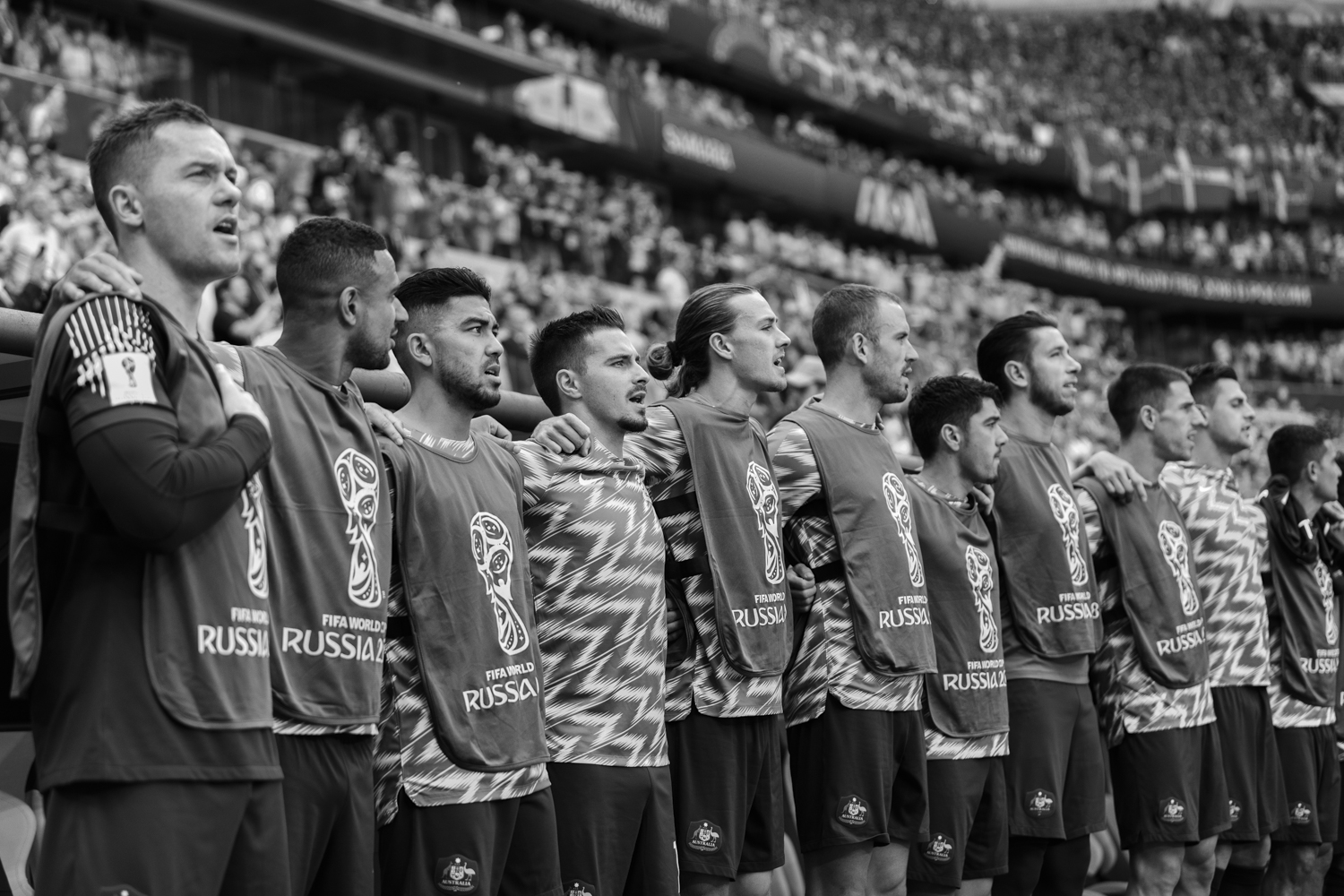 A group of soccer players stands shoulder-to-shoulder in black and white, wearing jerseys, in front of a blurred stadium crowd.