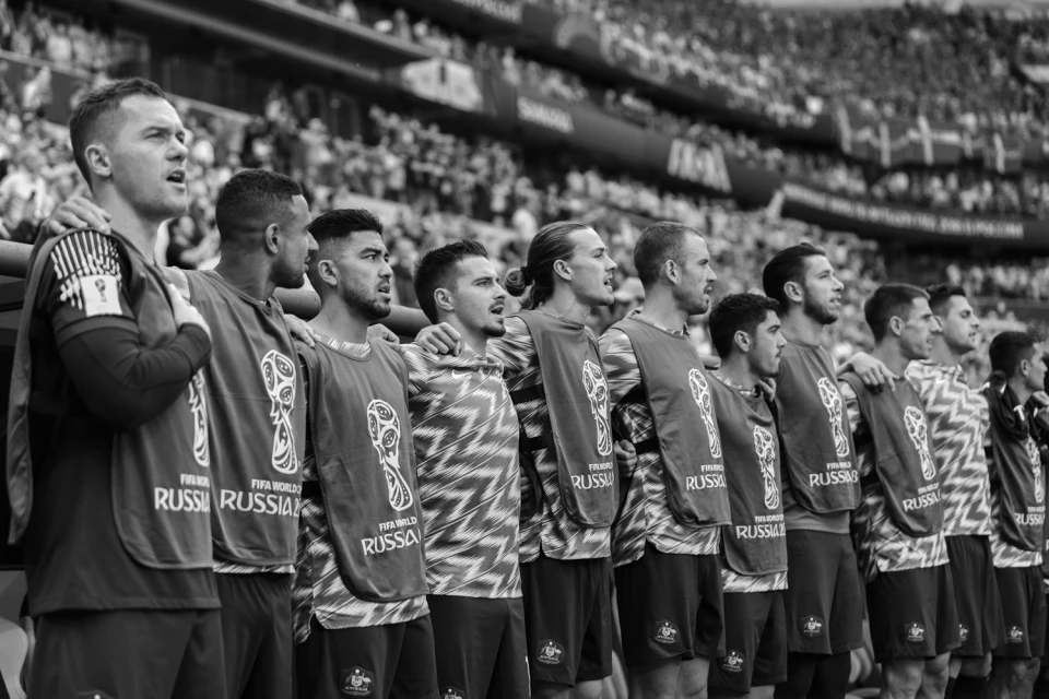 A group of soccer players stands shoulder-to-shoulder in black and white, wearing jerseys, in front of a blurred stadium crowd.