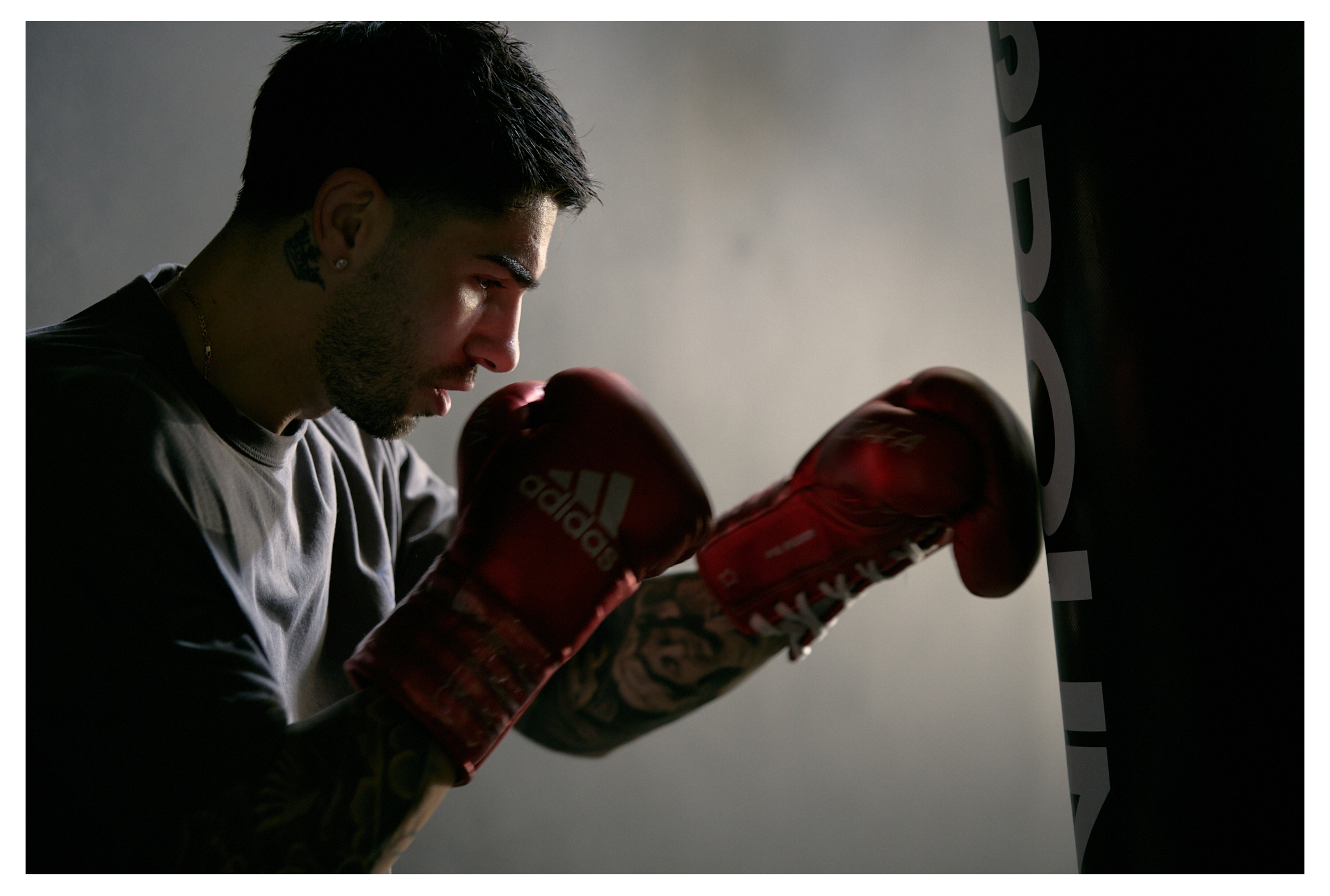 Boxer trains with red Adidas boxing gloves punching a black heavy bag in a dimly lit gym with a grey background and tattooed arms.