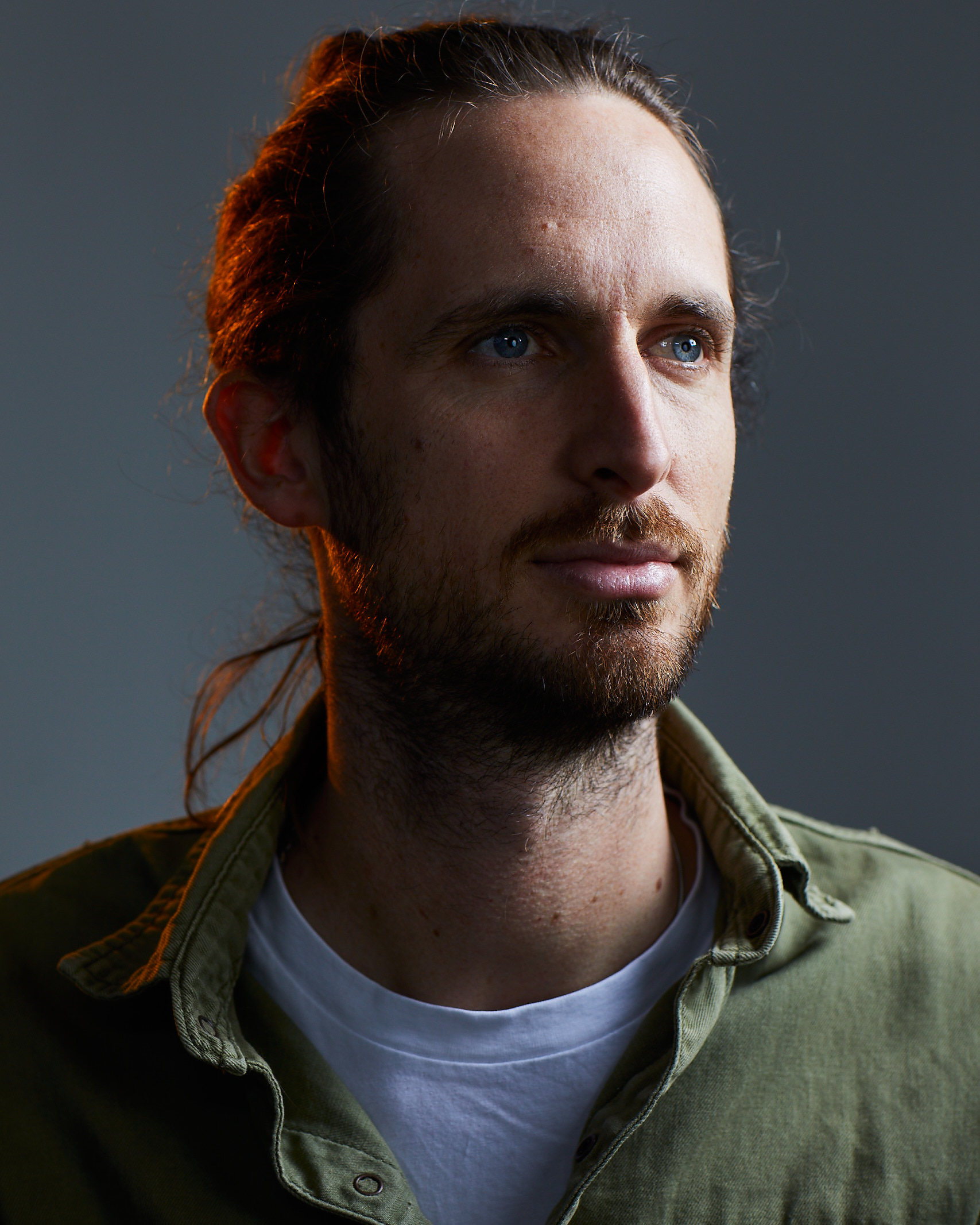 Portrait of a brunette adult male with tied back hair, light beard, wearing a green button-up shirt, and looking off-camera in a photo studio.