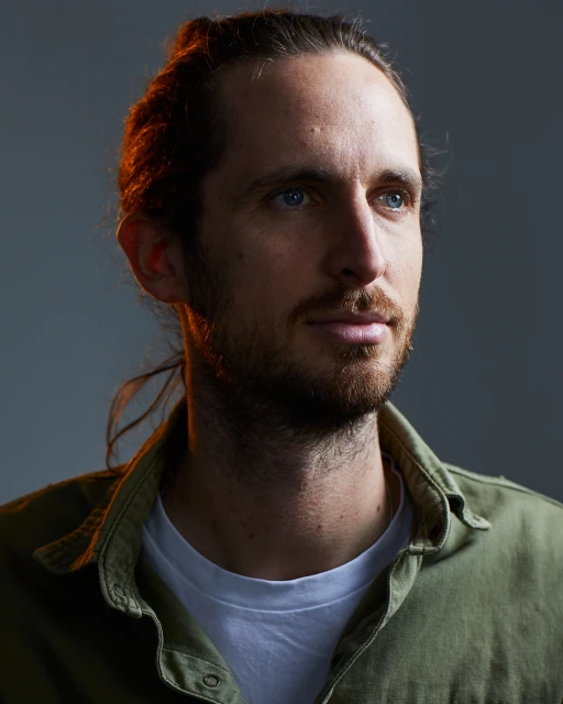 Portrait of a brunette adult male with tied back hair, light beard, wearing a green button-up shirt, and looking off-camera in a photo studio.