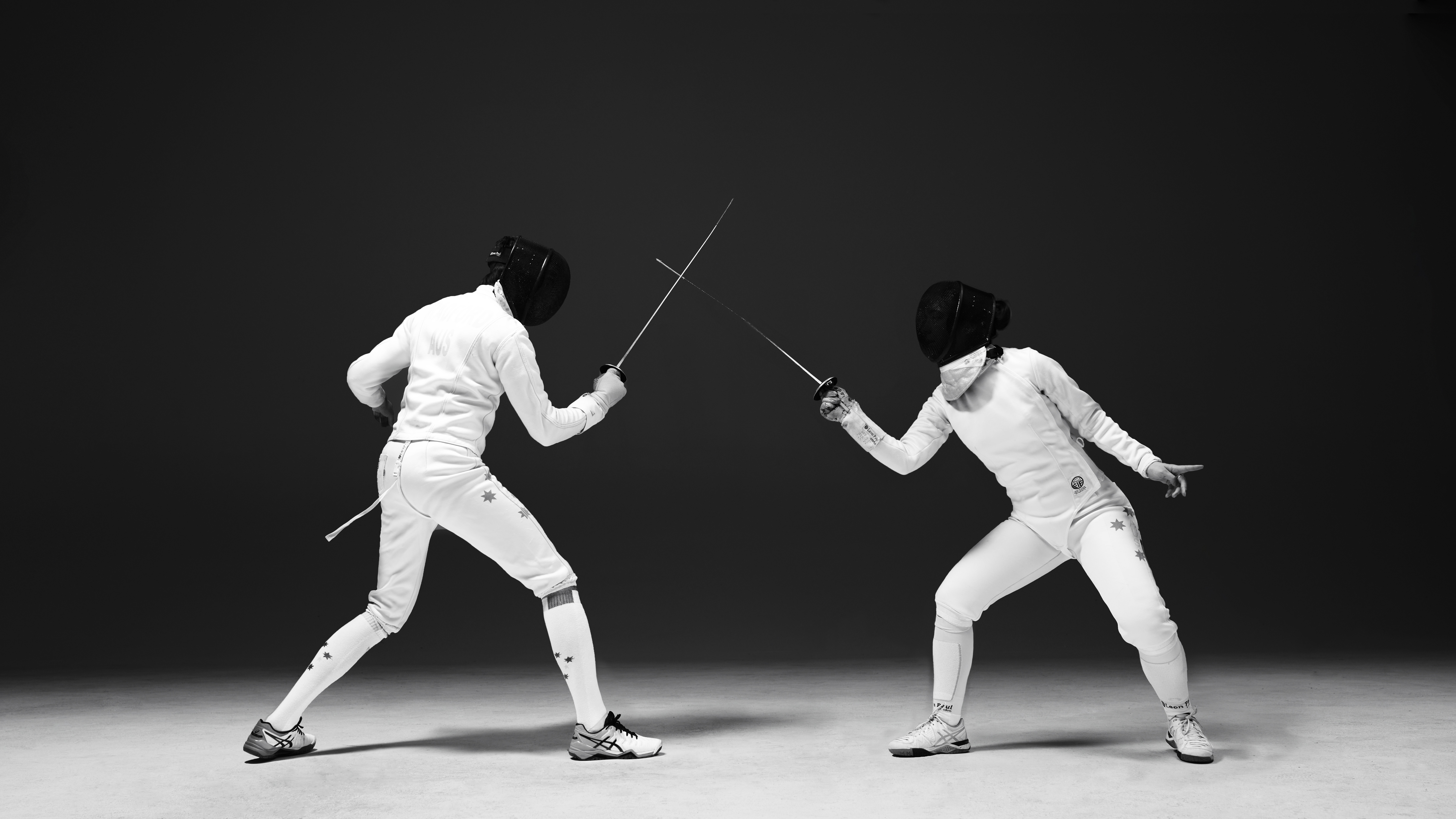 Two fencers in white uniforms and masks face off in a black and white action shot, swords crossed in a poised, competitive stance.
