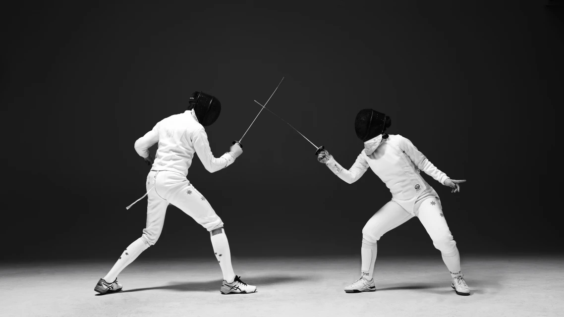 Two fencers in white uniforms and masks face off in a black and white action shot, swords crossed in a poised, competitive stance.