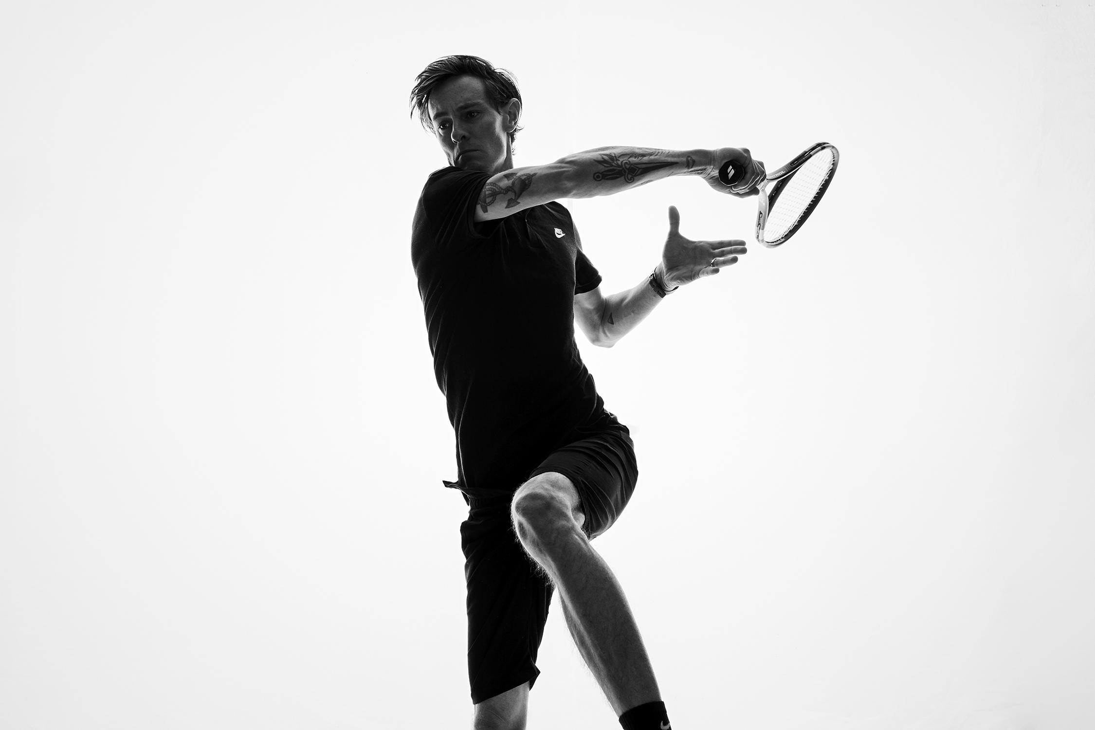 Monochrome shot of an adult male tennis player in motion, wearing a dark athletic outfit against a bright white backdrop.