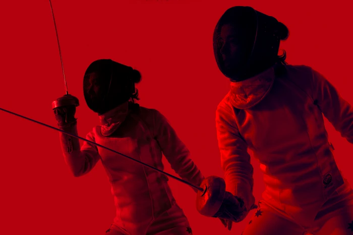 Two fencers in white protective gear and black masks holding foils against a vivid red background shot by Daniel Mazzarella.