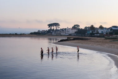 Five swimmers wearing swim caps and trunks wading into calm coastal waters along a sandy beach at dawn.