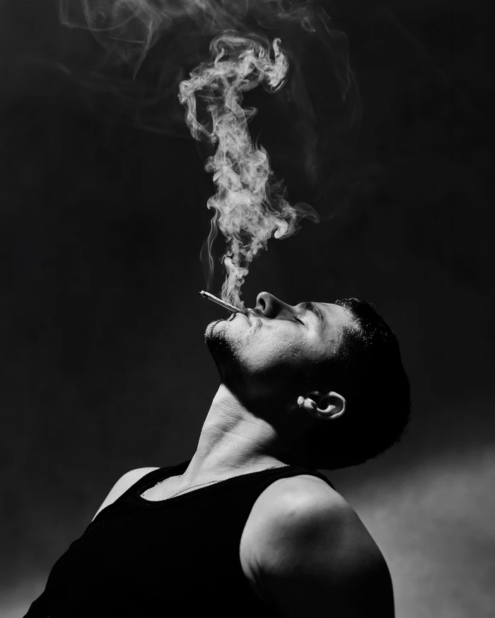 Monochrome portrait of a man in a black tank top smoking with intricate smoke trails rising against a dark background.