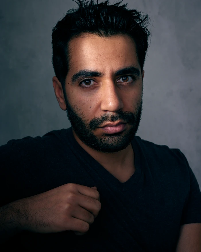 Close-up portrait of a bearded man in a dark v-neck t-shirt looking directly at the camera against a textured grey background.