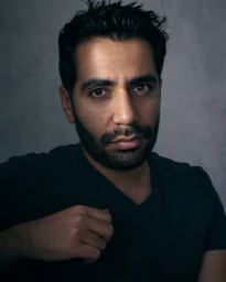 Close-up portrait of a bearded man in a dark v-neck t-shirt looking directly at the camera against a textured grey background.