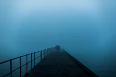 Silhouetted figures walk along a pier enveloped in dense blue fog with a metal railing and lone lamppost.