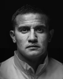 Close-up black and white portrait of a man wearing a high-collared zipped jacket against a dark background.